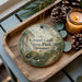 A decorative ornament with a scenic image of Grand Gulf State Park, Missouri, is displayed on a wooden tray alongside pine cones and a lit candle.