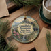 A decorative ornament with a scenic mountain landscape, featuring a bear, pine trees, and a waterfall, is displayed on a wooden surface alongside a cup of coffee and some papers.