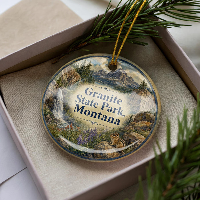 A decorative ornament in a box, featuring a scenic landscape with mountains, pine trees, and a waterfall.