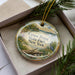 A decorative ornament in a box, featuring a scenic landscape with a lake, trees, and a bird.
