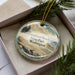 A circular ornament with a beach scene and text, placed in a box with pine branches.