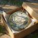 A decorative ornament in a wooden box, featuring a scenic landscape with mountains, waterfalls, and pine trees.