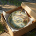 A circular ornament in a wooden box, featuring a scenic landscape with mountains, water, and wildlife.