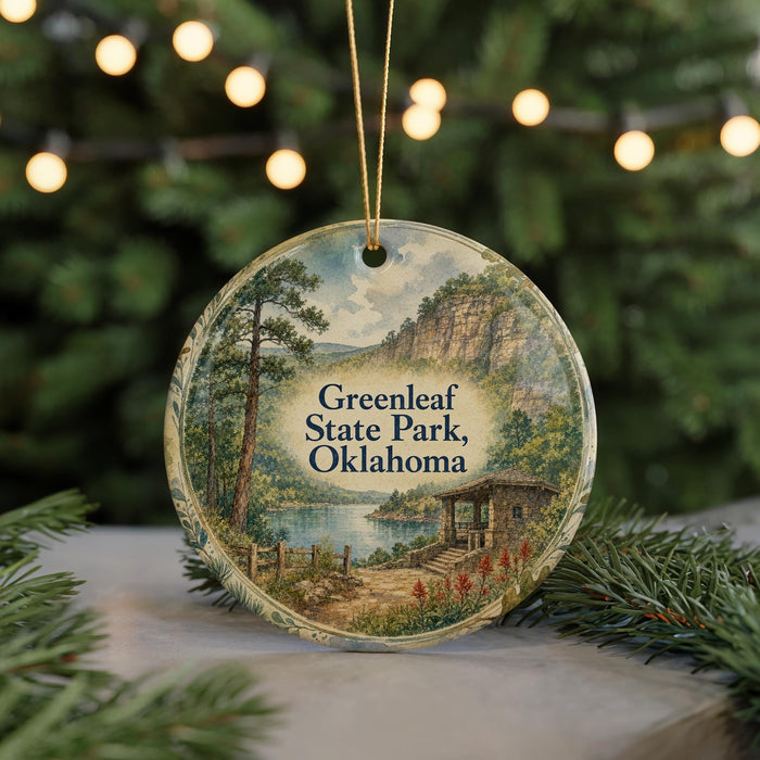 A circular ornament featuring a scenic landscape with a lake, mountains, and a cabin, hanging in front of a blurred Christmas tree with lights.