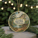 A circular ornament featuring a scenic landscape with a lake, mountains, and a cabin, hanging in front of a blurred Christmas tree with lights.