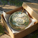 A decorative ornament in a wooden box, featuring a scenic landscape with a lake, trees, and a cabin.