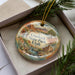 A decorative ornament in a box, featuring a scenic landscape with mountains, water, and greenery.