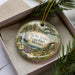 A decorative ornament in a box, featuring a scenic landscape with a castle, trees, and water.