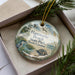 A decorative ornament in a box, featuring a scenic image of a beach and wildlife, including birds and a turtle.