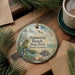 A round ornament with a beach scene and birds hangs on a wooden surface, accompanied by a cup of coffee and pine branches.