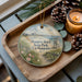 A decorative ornament featuring a beach scene with a lighthouse, pine cones, and a candle on a wooden tray.