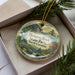A decorative ornament in a box, featuring a scenic landscape with a waterfall and pine trees.