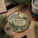 A round ornament with a scenic landscape of hanging rock, state park, and water, featuring pine trees and a waterfall, is displayed on a wooden surface alongside a cup of coffee and some papers.