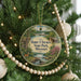 A round ornament hanging on a Christmas tree, featuring a scenic image of a river and a cabin surrounded by trees.