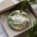 A decorative ornament with the text "Heavener Runestone State Park, Oklahoma" printed on it, placed inside a box with pine branches.