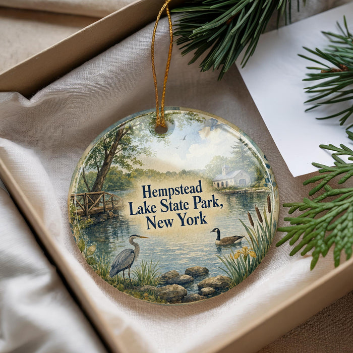 A decorative ornament with a scenic lake and park landscape, featuring a heron and a duck, is displayed in a box with pine branches.
