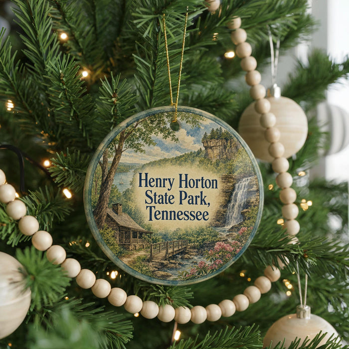 A round ornament hanging on a Christmas tree, featuring a scenic landscape with a waterfall and a cabin.