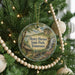 A round ornament hanging on a Christmas tree, featuring a scenic landscape with a waterfall and a cabin.