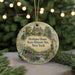 A decorative ornament featuring a historical scene with a house, a statue, and a bridge, surrounded by greenery and a string of lights.