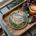 A decorative ornament featuring a scenic landscape of High Cliff State Park, Wisconsin, with pine cones and a lit candle on a wooden tray.