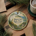A decorative ornament featuring a scenic landscape with cliffs, trees, and a lake, hanging on a wooden surface.