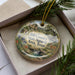 A decorative ornament in a box, featuring a scenic landscape with a deer, trees, and a waterfall.