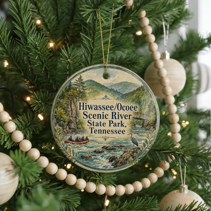 A round ornament hanging on a Christmas tree, featuring a scenic river and mountains in the background.