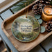 A wooden tray with a decorative ornament featuring a scenic image of Hochatown State Park, Oklahoma, along with pine cones and a lit candle.