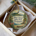 A decorative ornament in a box, featuring a scenic landscape with pine trees, a lake, and mountains.