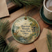 A decorative ornament featuring a scenic image of Huesston Woods State Park, Ohio, with a deer and a bridge in the background. The ornament is hanging on a wooden surface, surrounded by pine branches and a cup of coffee.