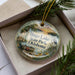 A decorative ornament with a scenic image of a lake and surrounding landscape, featuring a boat and a bird, is displayed in a box with pine branches.