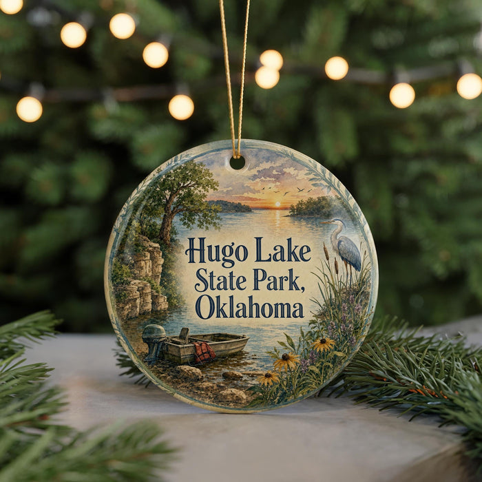 A round ornament with a painting of a lake and a bird, surrounded by greenery, hangs in front of a Christmas tree with lights.