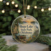 A round ornament with a painting of a lake and a bird, surrounded by greenery, hangs in front of a Christmas tree with lights.