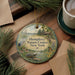 A round ornament with a nature scene hangs on a wooden surface, surrounded by pine branches and a cup of coffee.