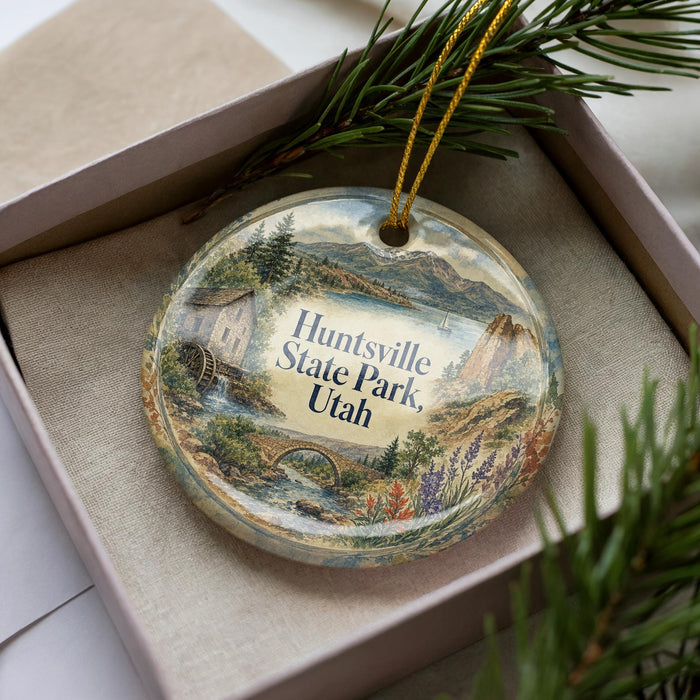 A decorative ornament in a box, featuring a scenic landscape with mountains, a river, and a small house.