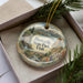 A decorative ornament in a box, featuring a scenic landscape with mountains, a river, and a small house.