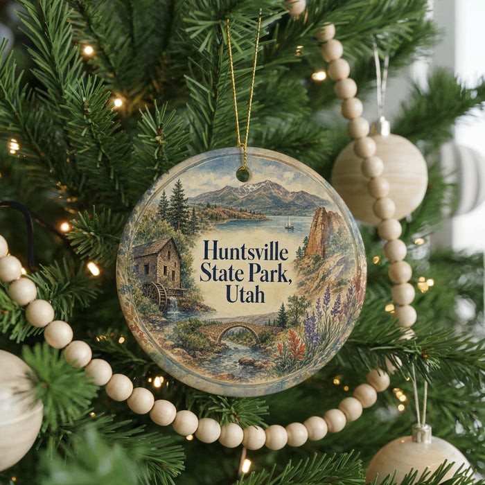 A round ornament hanging on a Christmas tree, featuring a scenic landscape with mountains, a river, and a cabin.
