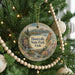 A round ornament hanging on a Christmas tree, featuring a scenic landscape with mountains, a river, and a cabin.