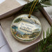 A decorative ornament with a landscape scene featuring mountains, pine trees, and a bird in flight, placed inside a box with a pine branch.