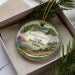 A decorative ornament with a scenic image of Independence Dam, a landmark in Ohio, is displayed in a box with pine branches.