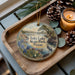 A decorative ornament with a scenic landscape of Inks Lake State Park, Texas, featuring a bird and surrounded by pine cones and a lit candle.