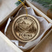 A circular ornament with a mountain landscape and text, placed in a box with a white tissue paper lining.