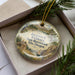 A decorative ornament with a scenic landscape, featuring a cabin, mountains, and a sailboat, is displayed in a box with pine branches.