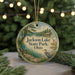 A round ornament with a scenic lake and forest landscape, featuring a bird and a cabin, is displayed on a table with Christmas lights in the background.