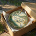 A round ornament in a box, featuring a scenic landscape with a lake, trees, and a bird, is displayed on a wooden surface.