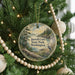 A circular ornament hanging on a Christmas tree, featuring a scenic landscape with a lake, trees, and a bird.
