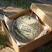 A circular ornament in a wooden box, featuring a scenic landscape with trees, a river, and a bird, is displayed on a wooden surface.