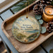 A decorative ornament featuring a beach scene with a lighthouse, pine cones, and a candle on a wooden tray.