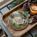 A wooden tray with a decorative ornament featuring a scenic landscape of Jenny Jump State Forest, New Jersey, along with pine cones and a lit candle.