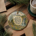 A round ornament with a scenic landscape featuring a waterfall, pine trees, and a meadow, hanging on a wooden surface.